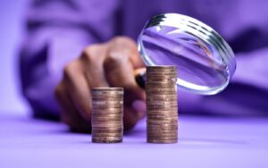 An analyst is holding a magnifying glass up to two piles of pennies, one stacked higher than the other, to represent the Disability Pay Gap. They are dressed in purple and the background and table in the image are purple to represent the Purple Pound, which is the term used to describe the spending power of disabled households.