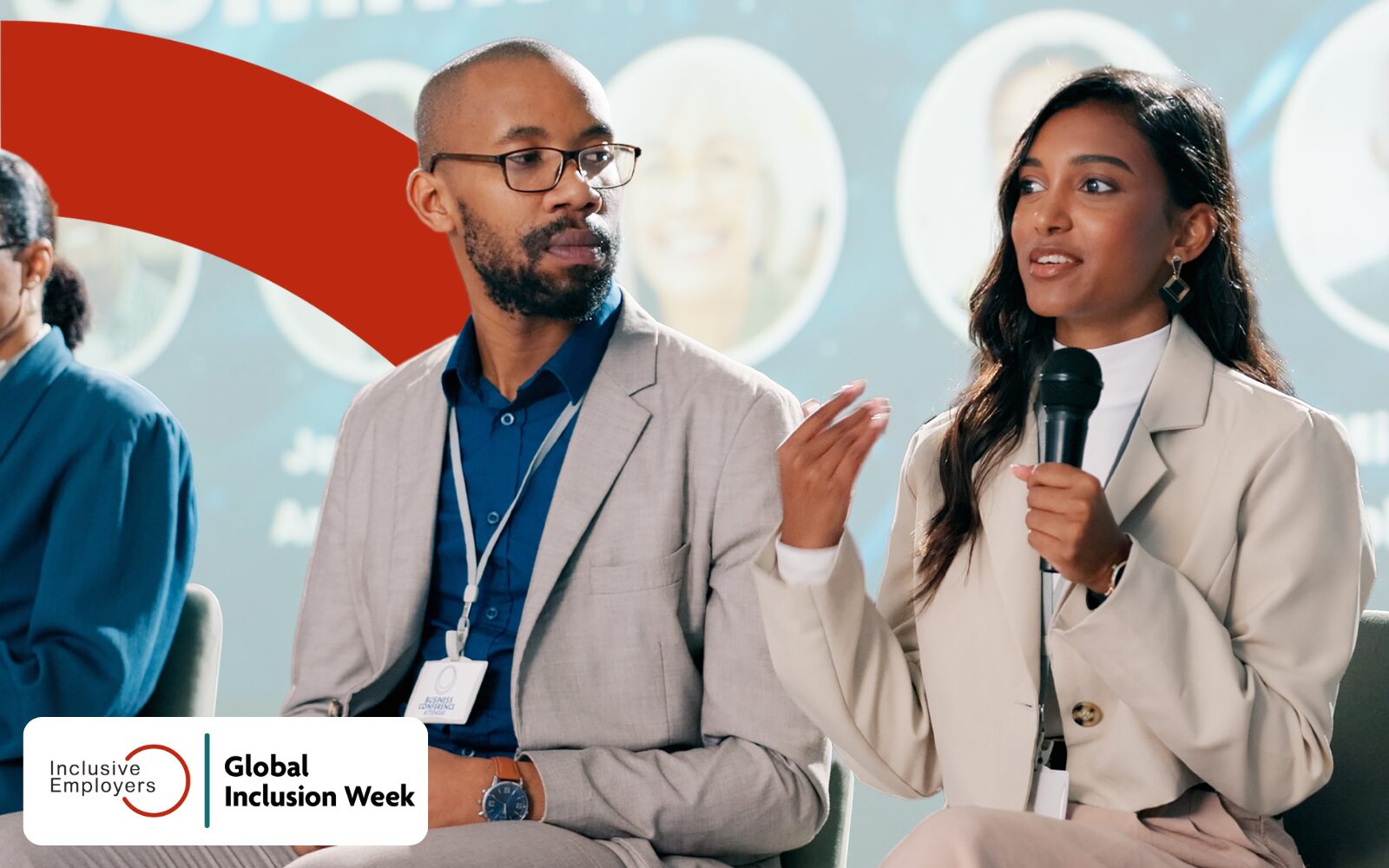 Photo of three people sitting in a row, only two are the focus of the image. One is a man, one is a woman and they are both wearing light coloured suits. The woman is gesturing with one hand while holding a microphone as the man looks at her and listens.