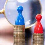 An office worker holds a looking glass up to two figurines. Figurine one is blue and is standing on a higher stack of coins than figurine two, a red figurine standing on a smaller pile of coins. This image is supposed to represent pay gaps between people with two different characteristics.