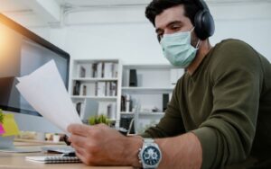 Man wearing a medical facemask whilst reading paperwork at his homebased work station.