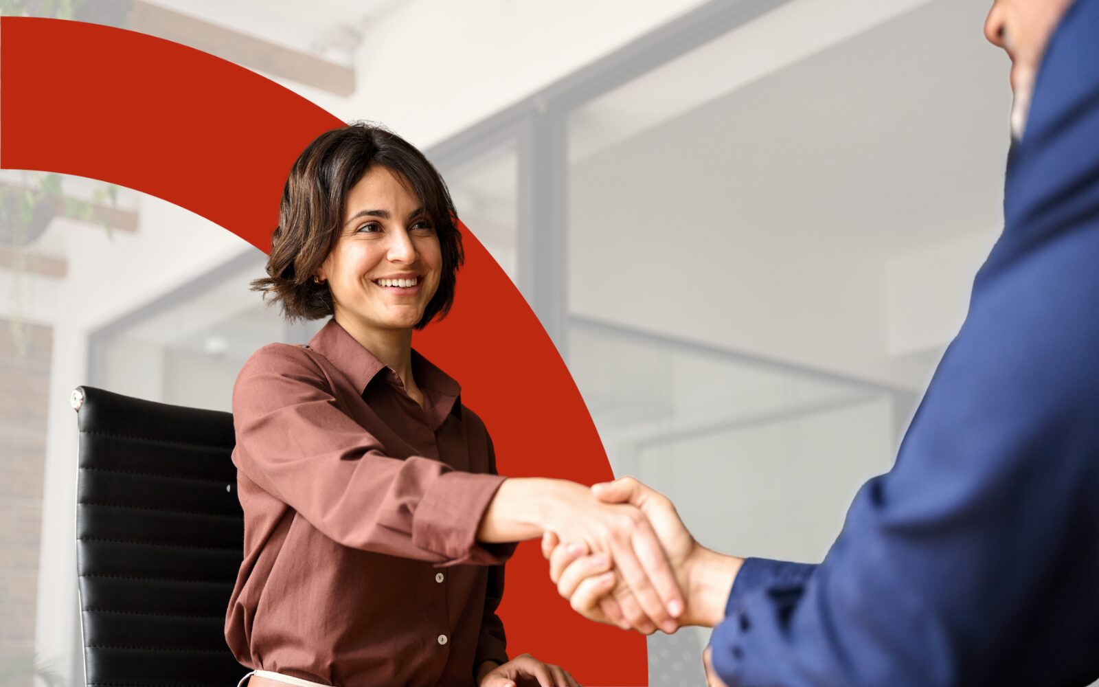 Photo of a smiling woman with a short brown bob haircut, wearing a red-brown shirt. She is shaking hands with someone in a blue shirt slightly off camera.