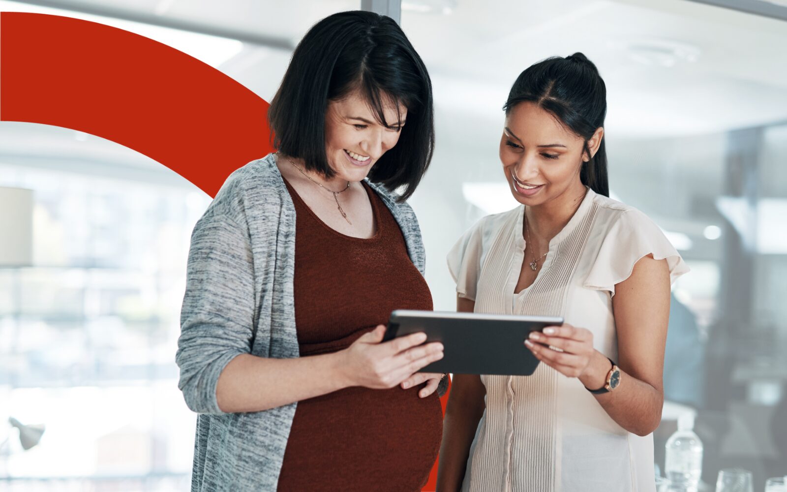 Photo of two women looking down at a tablet. They are both smiling and wearing smart casual outfits that are white, grey and red. The women on the left is visibly pregnant and has her hand on her baby bump.
