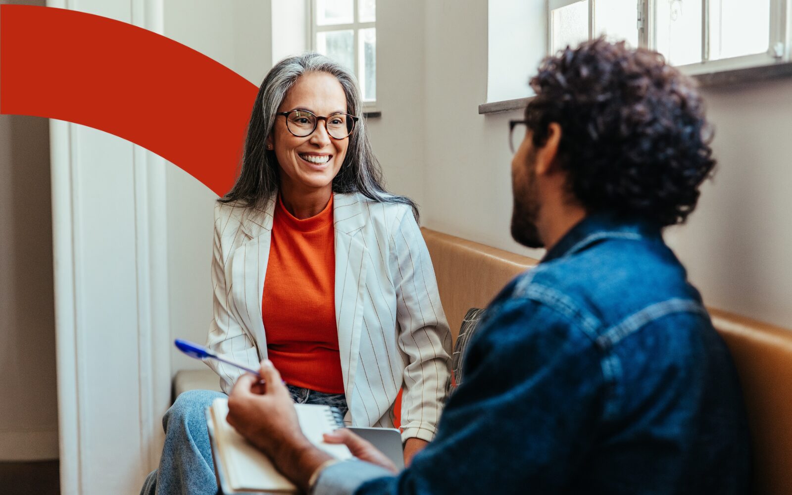 An image of two individuals sitting facing each other and smiling. One has a grey jacket, red shirt and long hair. The other has a blue jacket and short black hair.