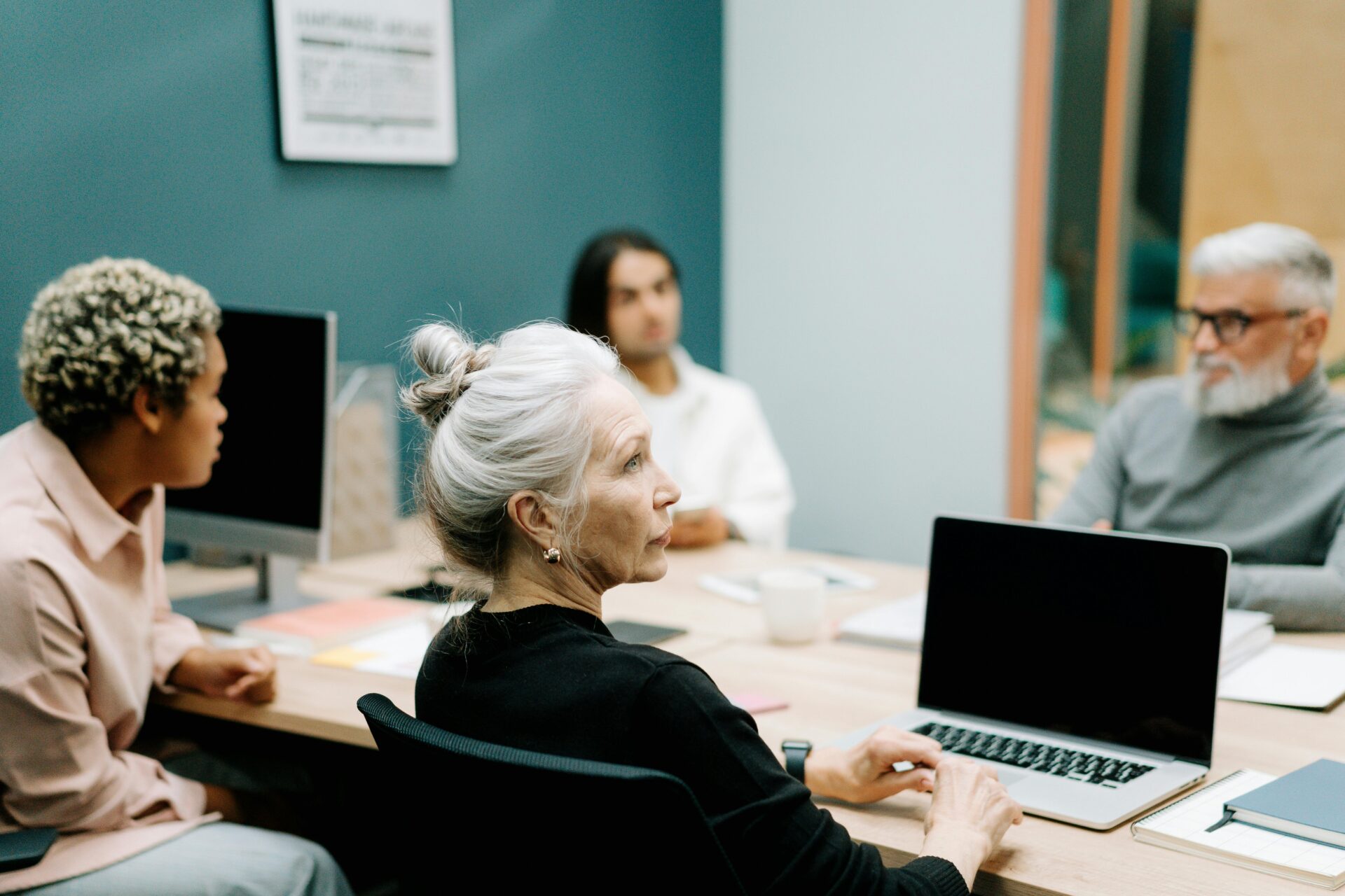 Photo of four people sitting around a table, the main person in the centre of the photo is a woman with silver and white hair and a black shirt who is looking off screen.