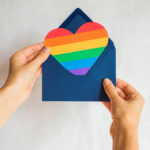 A pair of hands open a dark blue envelope and take out a rainbow heart