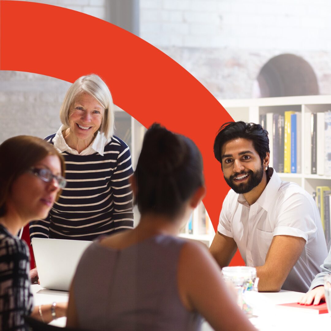 Photo shows a mature, feminine person and south-east Asian masculine person in a meeting with co-workers. A red arch intersects the image.